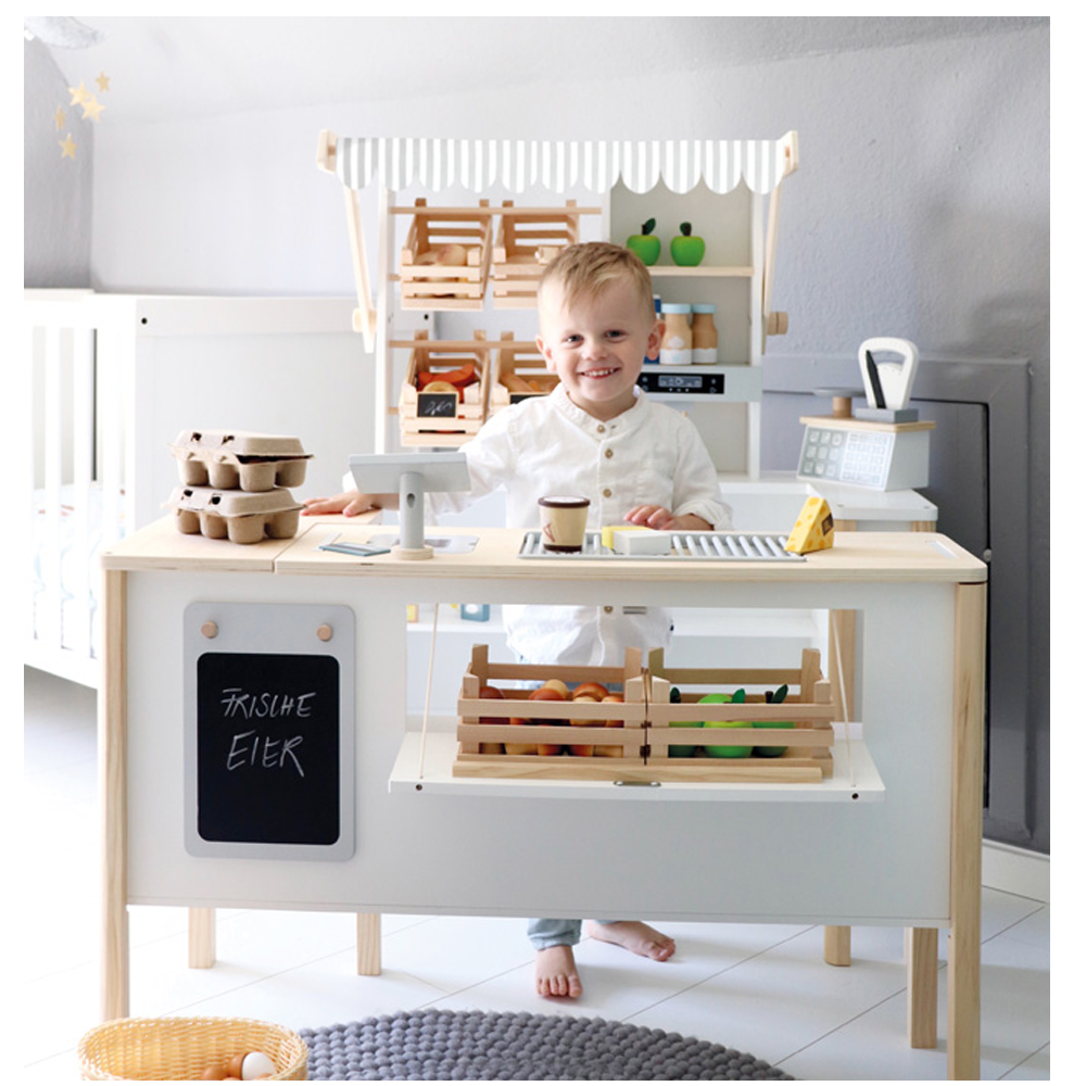 Hinter dem Kaufladen und der Kinderküche fresh spielt ein kleines Kind den Ladenbesitzer, mit Holzkisten und einer Spielkasse.