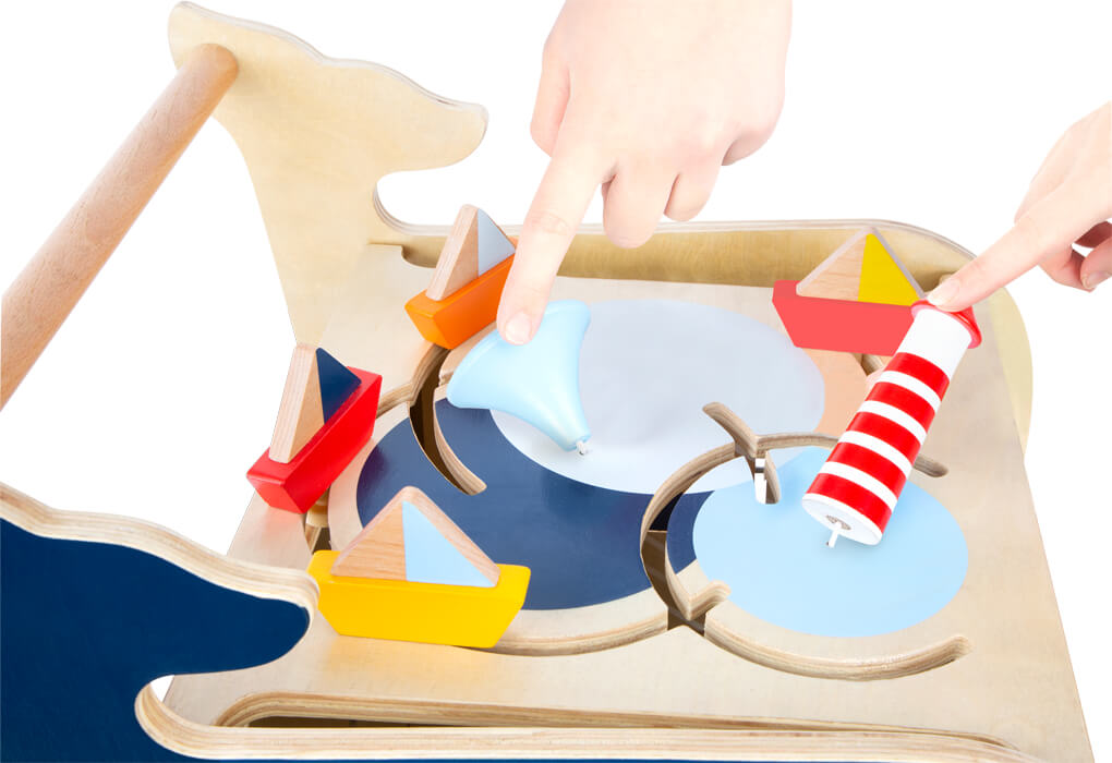 Zwei Hände bewegen bunte Holzboote und einen Leuchtturm auf dem Lauflernwagen Wal, einem hölzernen Aktivitätsspielzeug zum Thema Wasser.