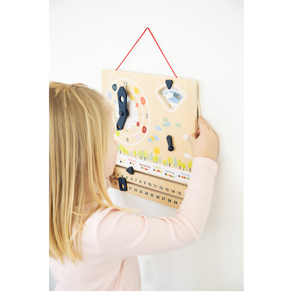 Ein junges Mädchen stellt die Lernuhr mit Kalender „Educate“ ein, eine hölzerne Lernuhr mit Kalender, die an einer weißen Wand hängt.