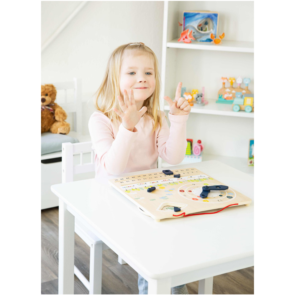 Ein junges Mädchen sitzt lächelnd an einem Tisch und hält sechs Finger hoch. Vor ihr liegt das Spiel „Educate“ mit Lernuhr und Kalender.