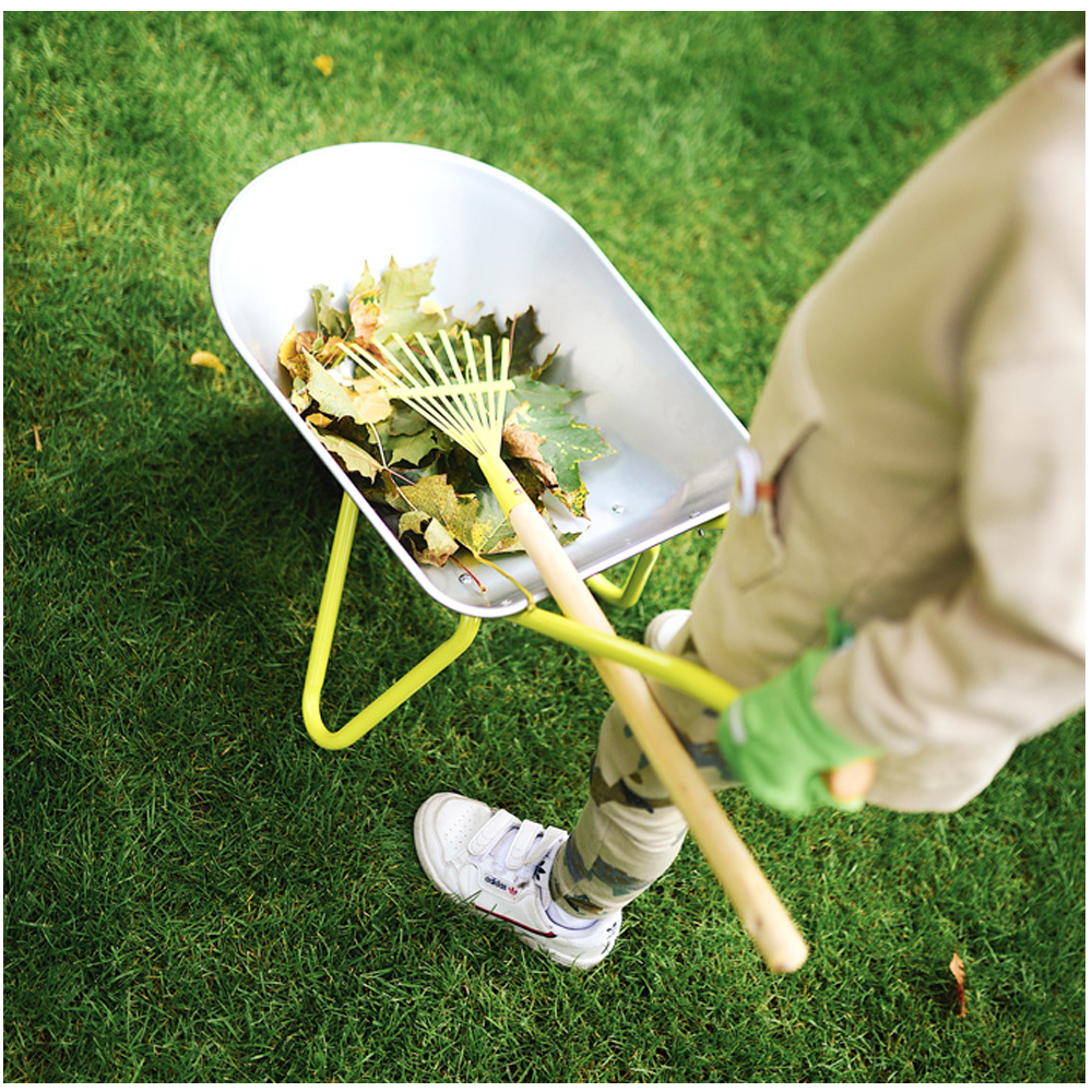 Ein Kind steht auf grünem Gras neben einer Schubkarre mit Gartenwerkzeug und hält einen Rechen mit Blättern, die in der Schubkarre aufgehäuft sind.