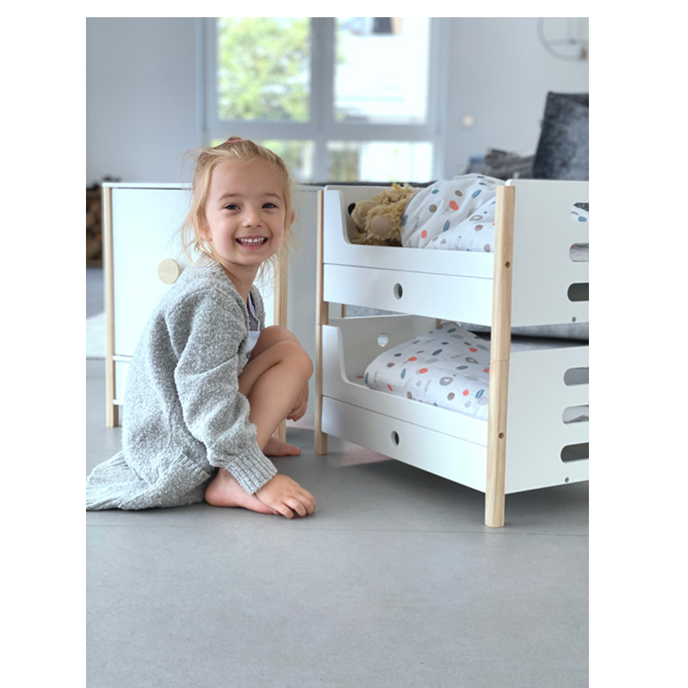 Ein lächelndes junges Mädchen kniet in einem hellen Zimmer neben dem Puppenhochbett „Little Button“, einem weißen Kinderbett mit gemusterter Bettwäsche.