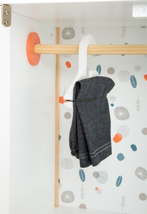 Im Puppenschrank „Little Button“ mit gemusterter Tapete hängt eine dunkle Jeans auf einem weißen Kleiderbügel.