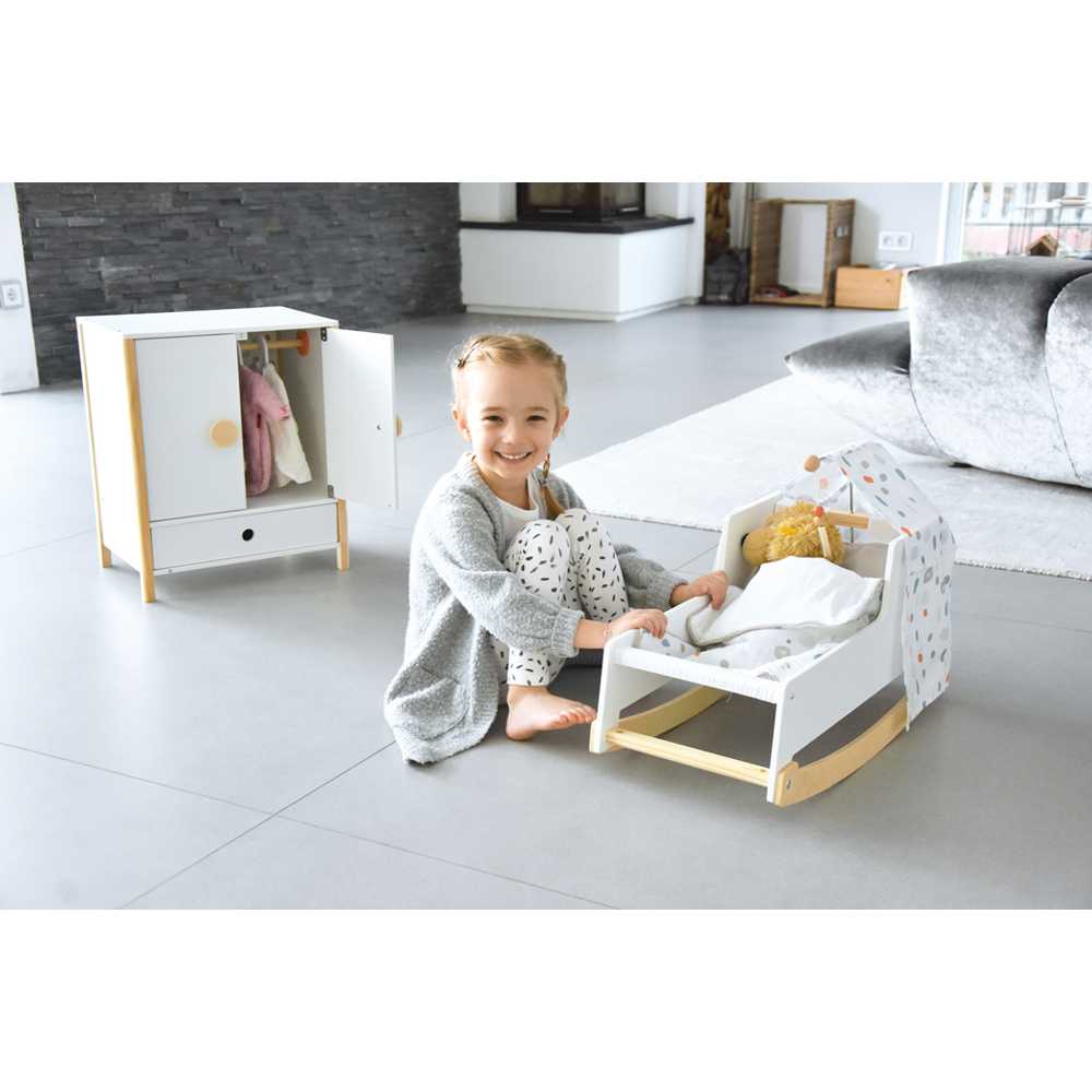 Ein lächelndes Mädchen sitzt auf dem Boden neben einem Spielzeugbett mit einer Puppe und dem weißen Puppenschrank „Little Button“ in der Nähe.