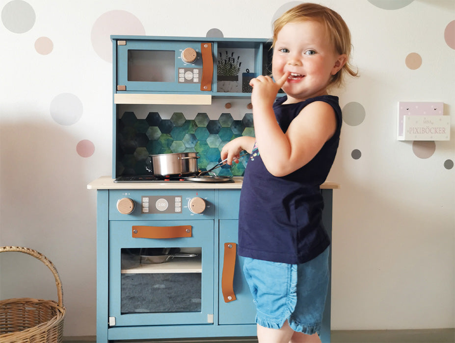 Ein kleines Kind lächelt, während es mit dem „leckeren“ Spielküchenset von Kinderküche spielt und einen Löffel an den Mund hält.