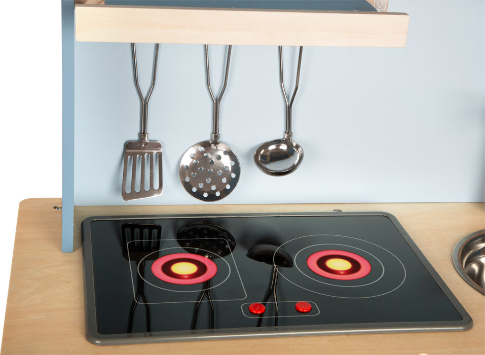 Die Kinderküche „tasty“ ist ein Spielzeugküchenherd mit roten Kochplatten, zwei Knöpfen und vier darüber hängenden Metallutensilien vor einer blauen Wand.