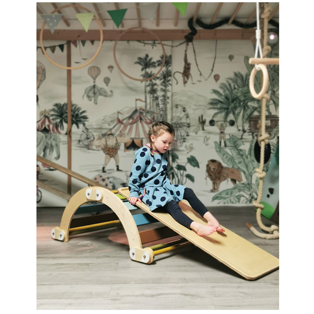 Ein junges Mädchen in einem blauen gepunkteten Kleid rutscht in einem Spielzimmer mit Zirkusthema die hölzerne Indoor-Rampe Rutschbrett 3 in 1 „Adventure“ hinunter.