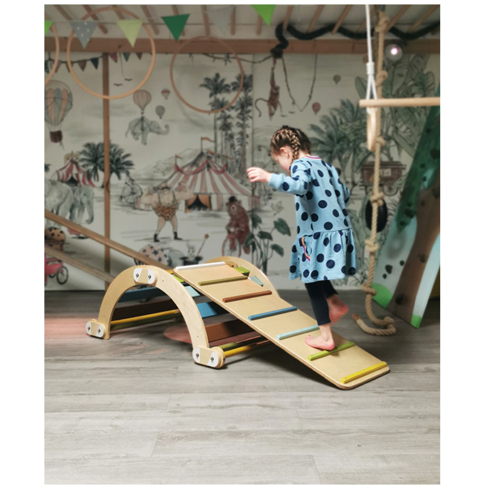 Ein junges Mädchen in einem blauen gepunkteten Kleid spielt barfuß im Haus auf dem Holzkletterspielzeug Rutschbrett 3 in 1 „Adventure“.