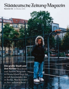 Ein Kind in einem Kapuzenpulli steht auf einem nassen Basketballplatz vor den Gebäuden der Stadt, unter einem bewölkten Himmel.