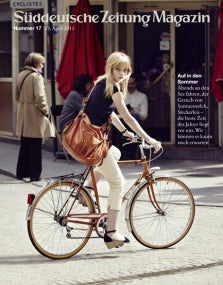Eine Frau mit einer braunen Tasche fährt mit dem Fahrrad auf einer städtischen Straße, mit roten Regenschirmen und Menschen im Hintergrund.