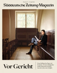 Ein Mann sitzt in einem leeren Gerichtssaal mit Holzbänken und großen Fenstern und liest auf dem Cover einer Zeitschrift.
