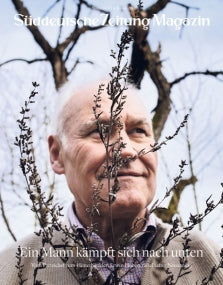 Älterer Mann, der hinter trockenen Ästen lächelt, mit Bäumen und bewölktem Himmel im Hintergrund. Text auf der Titelseite der Zeitschrift sichtbar.