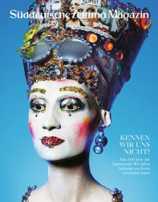 Eine Person mit dramatischem Make-up und einem farbenfrohen, kunstvollen Kopfschmuck posiert vor einem blauen Hintergrund.