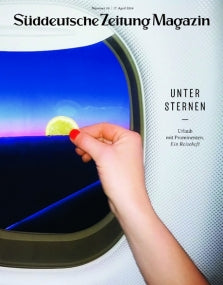 Eine Hand hält eine Limettenscheibe gegen ein Flugzeugfenster, das einen blauen Himmel und einen Sonnenuntergang zeigt.