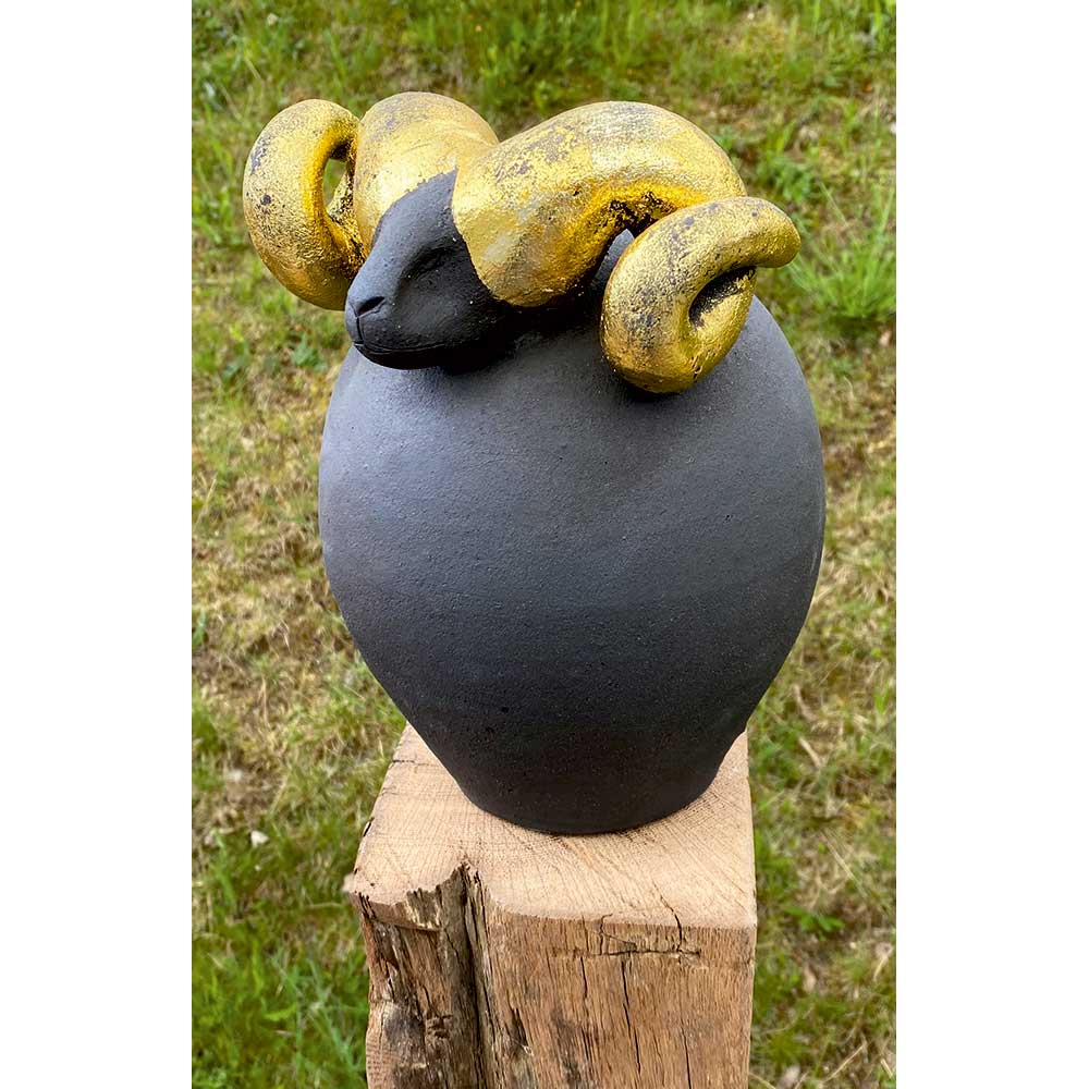 Schwarze Widderskulptur aus Keramik mit gelben Hörnern, im Freien auf einem Holzblock mit Gras im Hintergrund ausgestellt.