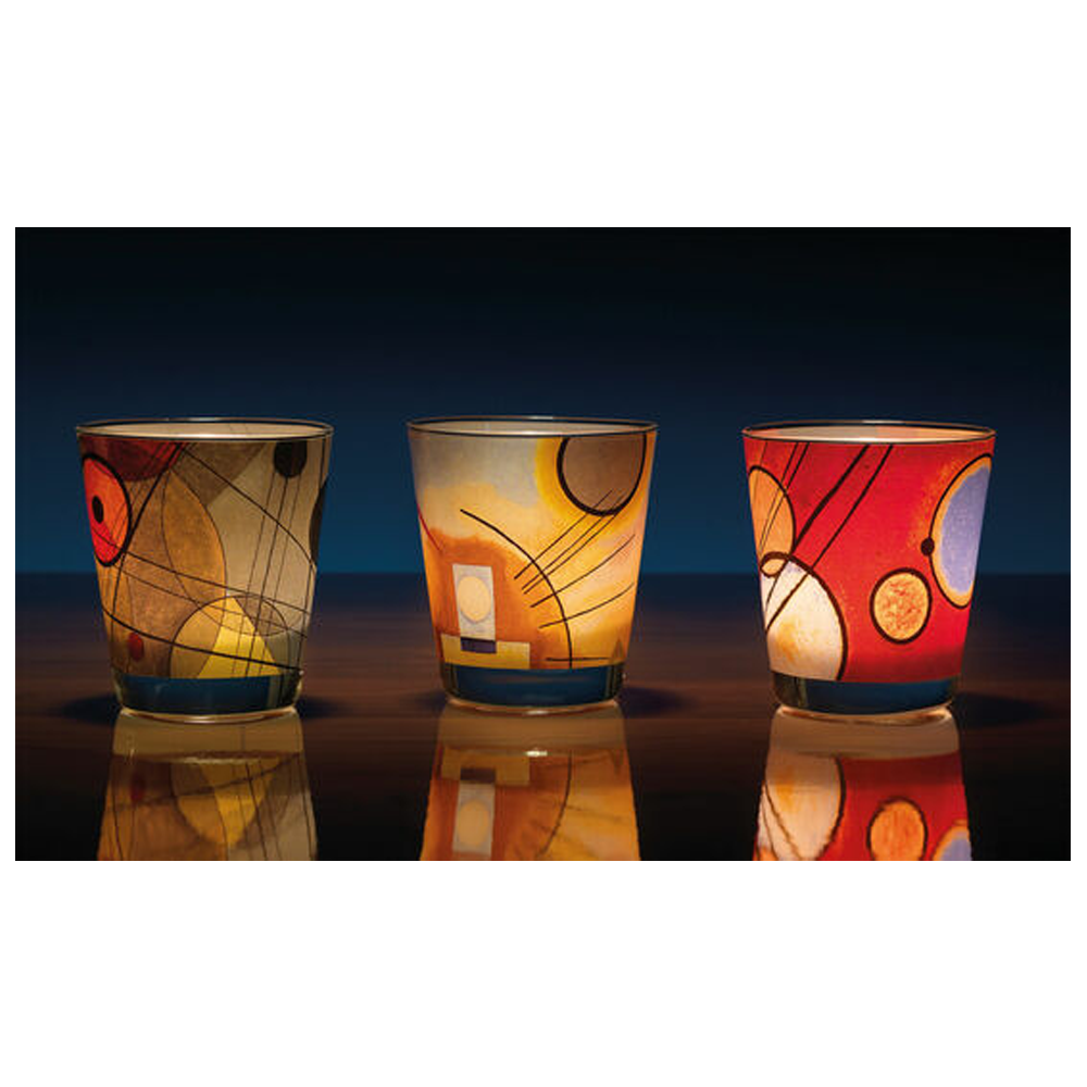 Drei bunte, gemusterte Kerzenhalter aus Glas leuchten von innen und reflektieren das Licht auf einer glänzenden Oberfläche.