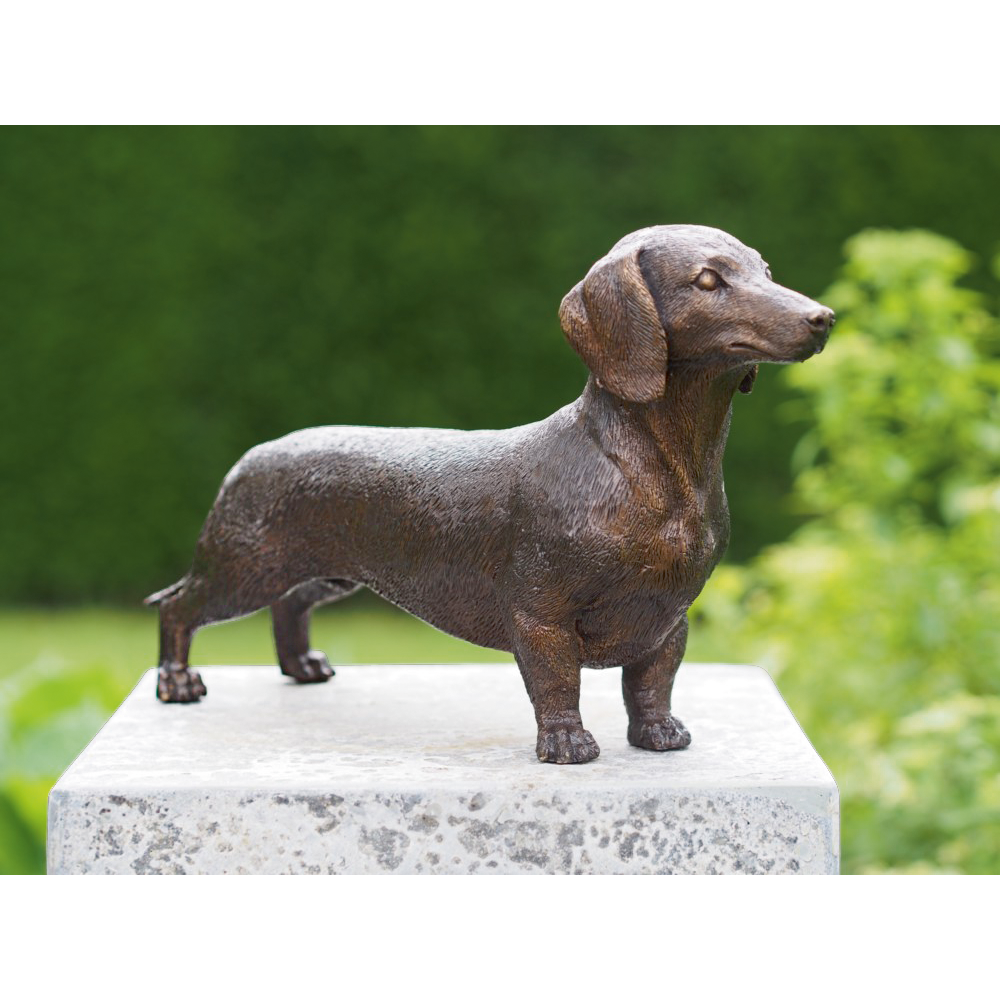 Bronzene Dackelstatue auf einem Steinsockel mit viel Grün im Hintergrund.