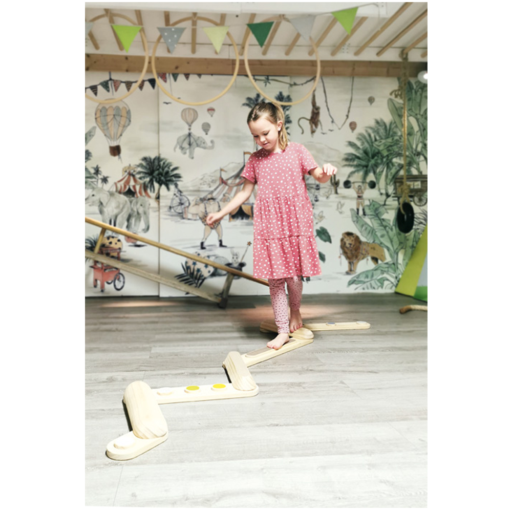 Ein junges Mädchen in einem rosa Kleid balanciert auf dem Holzspielzeug „Balancierstege Adventure“ im Innenbereich, im Hintergrund ein buntes Wandgemälde.