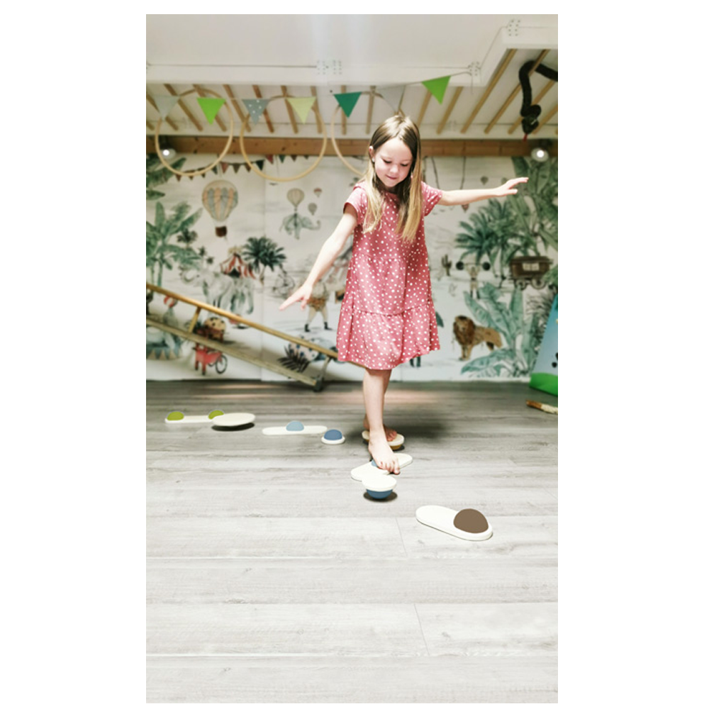 Ein junges Mädchen in einem rosa Kleid balanciert auf den Balanciersteinen „Adventure“ in einem hellen, verspielten Innenraum mit einer Wandmalerei.