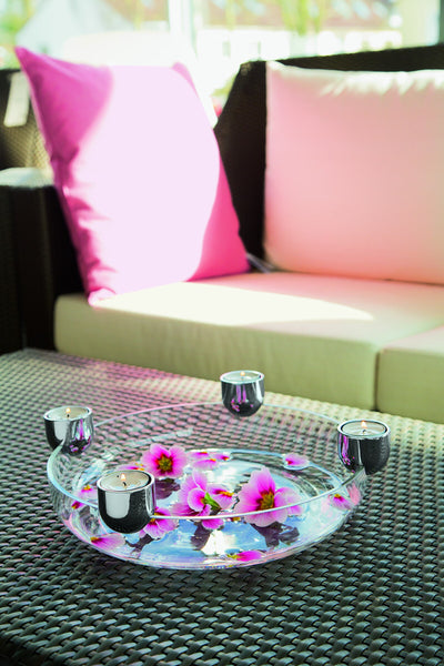 Tablett mit schwimmenden Blumen und Teelichtkerzen auf einem Korbtisch, rosa Kissen auf einem Sofa im Hintergrund.