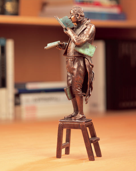 Bronzestatue eines Mannes, der ein Buch liest, während er auf einer kleinen hölzernen Trittleiter steht und weitere Bücher hält.