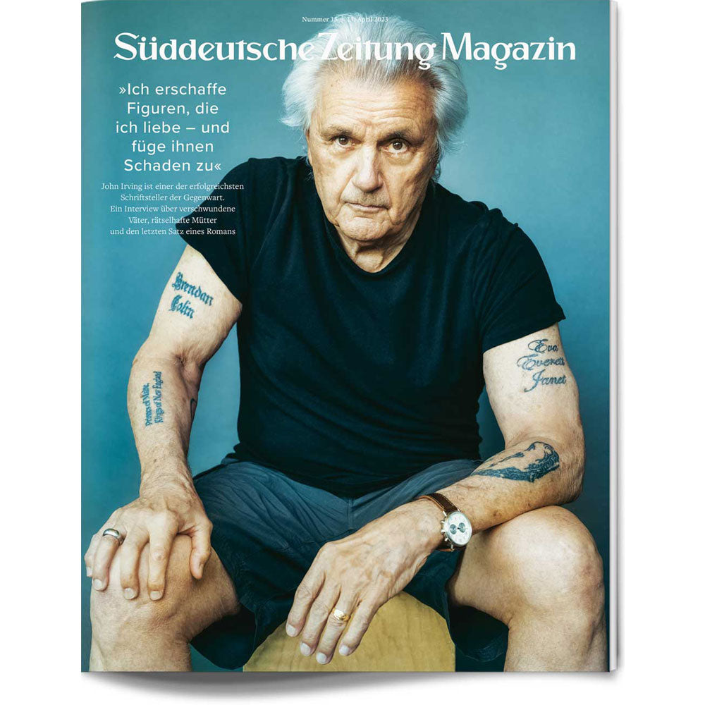 Älterer Mann mit grauen Haaren sitzt auf einem Hocker vor einer blauen Wand, trägt ein schwarzes Hemd und Shorts und zeigt tätowierte Arme.