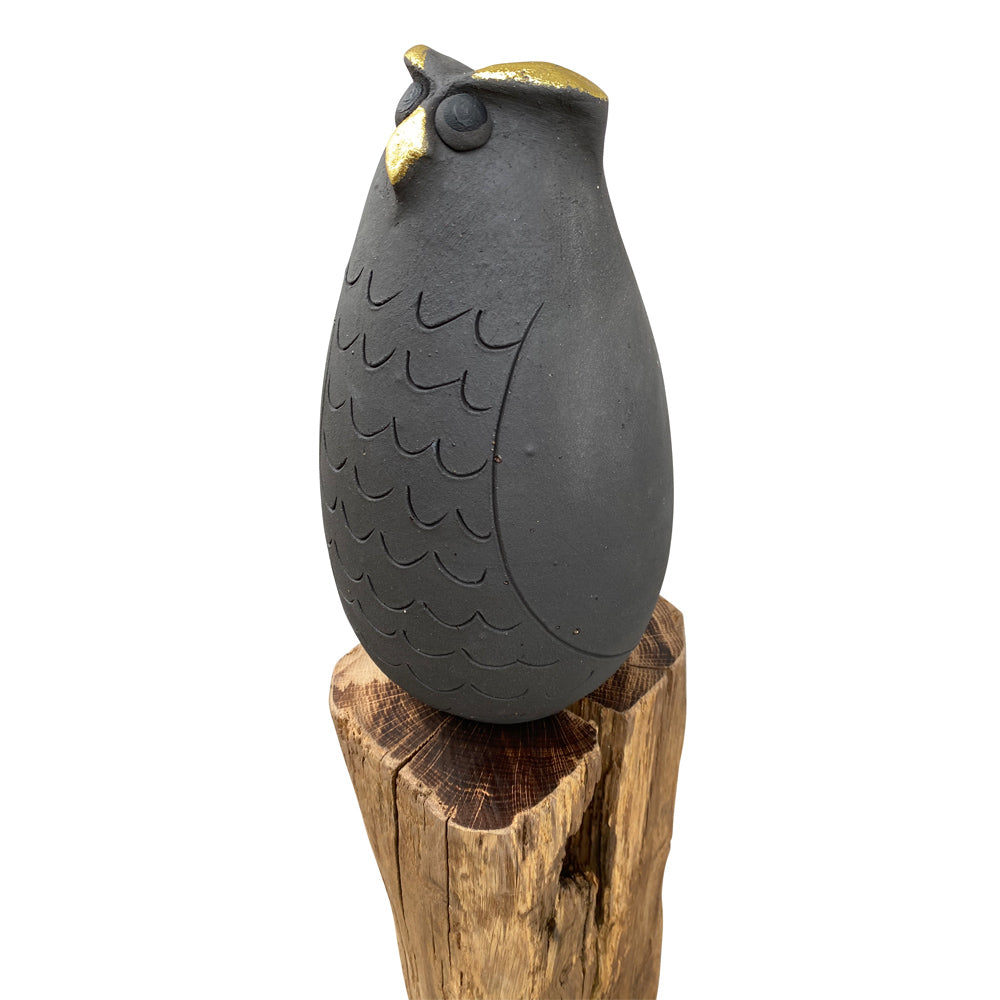Schwarze Eulenskulptur mit goldenem Schnabel, thront auf einem groben Holzstumpf vor einem weißen Hintergrund.