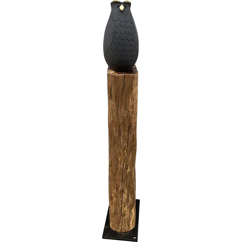Eine schwarze Eulenskulptur mit gelben Augen sitzt auf einem hohen Holzpfosten mit schwarzem Sockel.