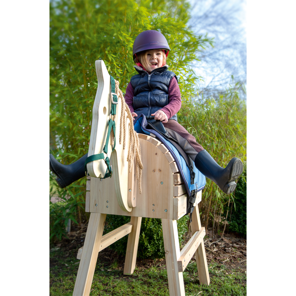 Ein lächelndes Kind mit Helm und Stiefeln sitzt draußen auf einem Holzpferd-Spielpferd in der Nähe grüner Bäume.