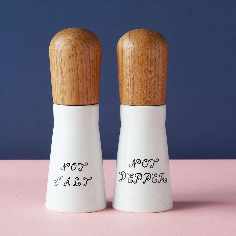 Das Gewürzmühlen-Set „Not Salt, Not Pepper“ besteht aus zwei weißen Streuern mit Holzdeckeln mit der Aufschrift „Not Salt“ und „Not Pepper“, abgebildet auf rosa-blauem Hintergrund.