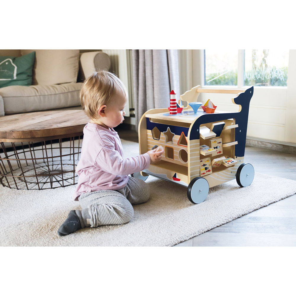 Ein Kleinkind spielt in einem gemütlichen Wohnzimmer mit dem Lauflernwagen Wal, einem hölzernen Spielzeugwagen zum Formensortieren.