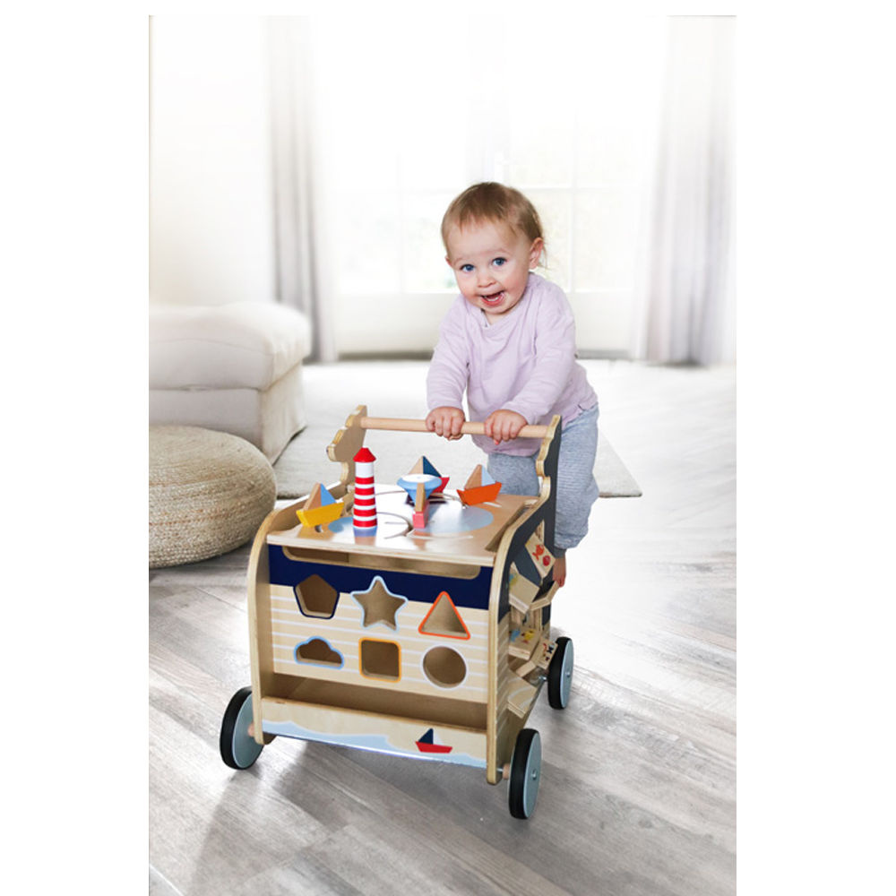 Ein lächelndes Baby steht mit dem Lauflernwagen Wal aus Holz in einem hellen, modernen Wohnzimmer.