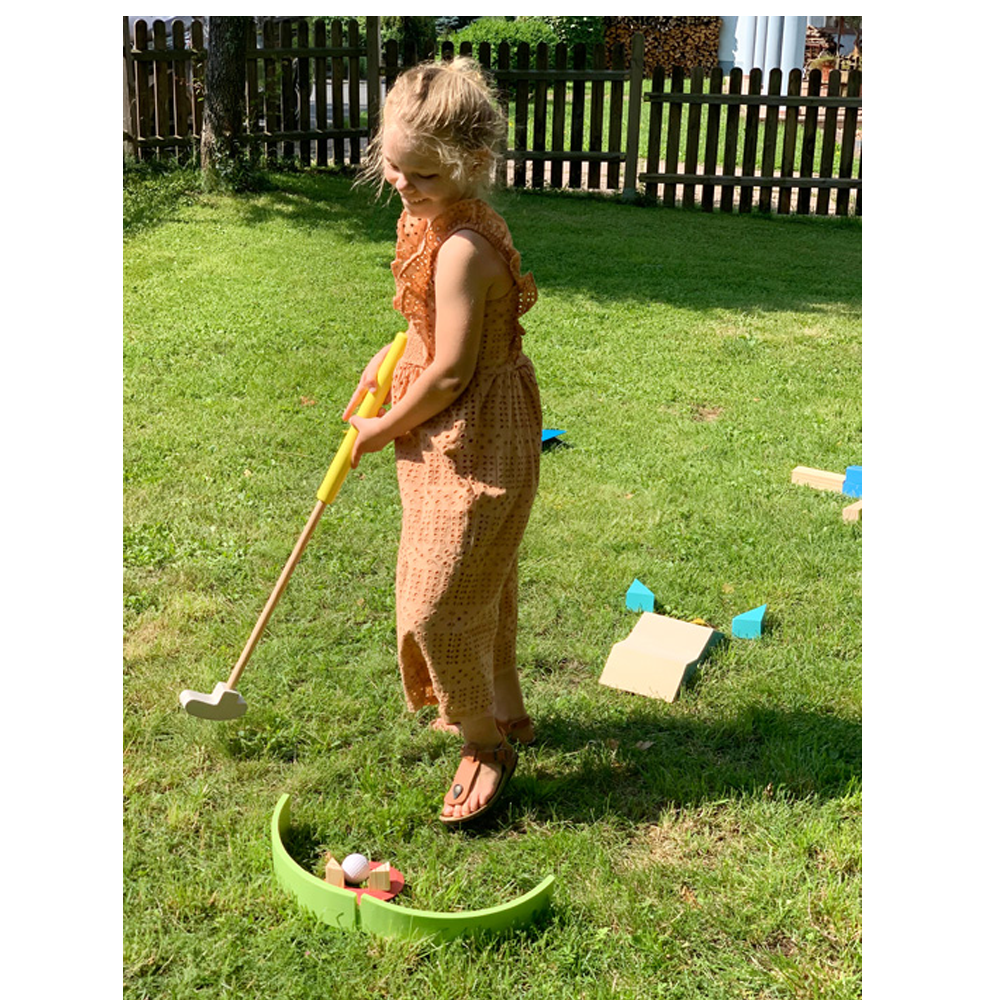 Ein junges Mädchen im braunen Kleid spielt Minigolf auf grünem Rasen mit dem Minigolf-Set „Active“ und hält einen gelb-weißen Schläger in der Hand.