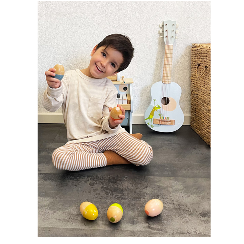 Ein lächelndes Kind sitzt auf dem Boden und hält Rhythmusspielzeuge „Groovy Beats“ von Musikeier in der Hand, im Hintergrund sind eine kleine Gitarre und ein Weidenkorb zu sehen.