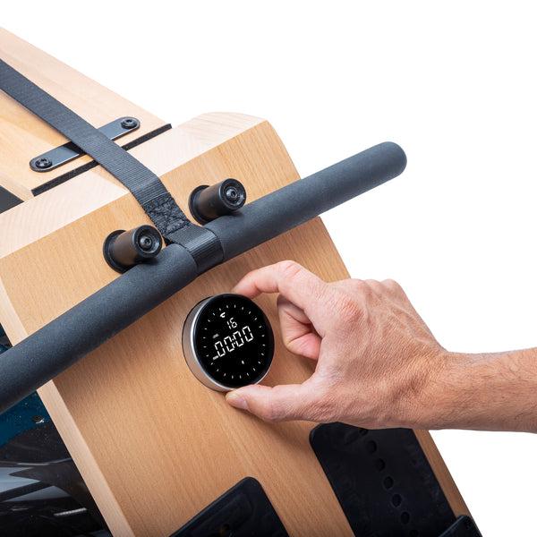 Eine Hand stellt eine runde Digitalanzeige auf einem hölzernen Rudergerät vor einem weißen Hintergrund ein.