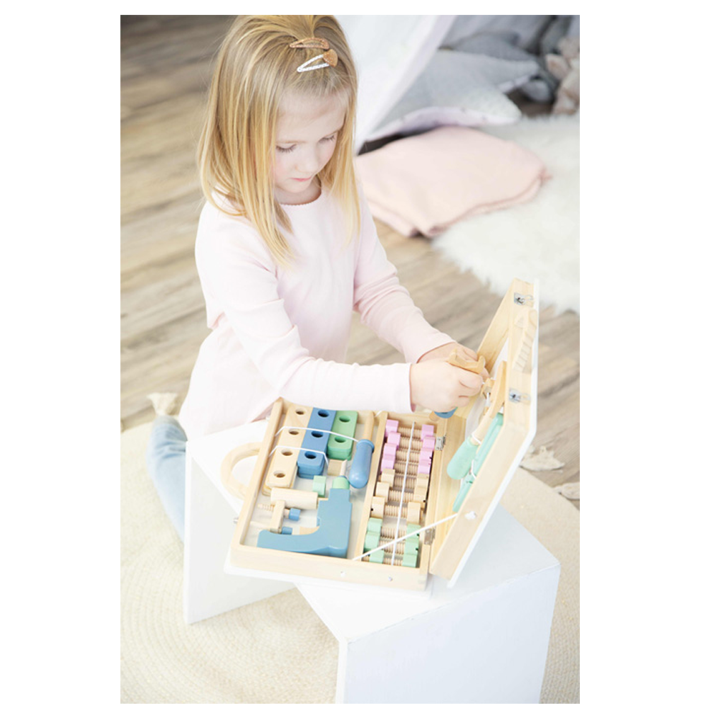 Ein junges Mädchen spielt mit dem Werkzeugkoffer „Nordic“, einem hölzernen Spielzeug-Werkzeugkasten auf einem weißen Würfel in einem gemütlichen Raum.
