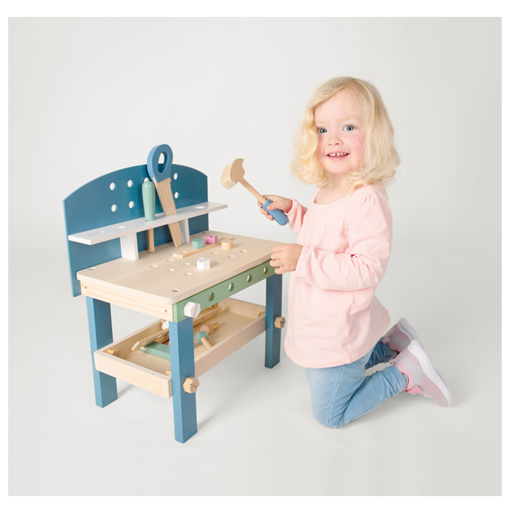 Ein kleines Kind lächelt in die Kamera, während es kniet und mit der Werkbank kompakt „Nordic“ spielt, einer Spielzeugwerkbank aus Holz mit Werkzeugen.