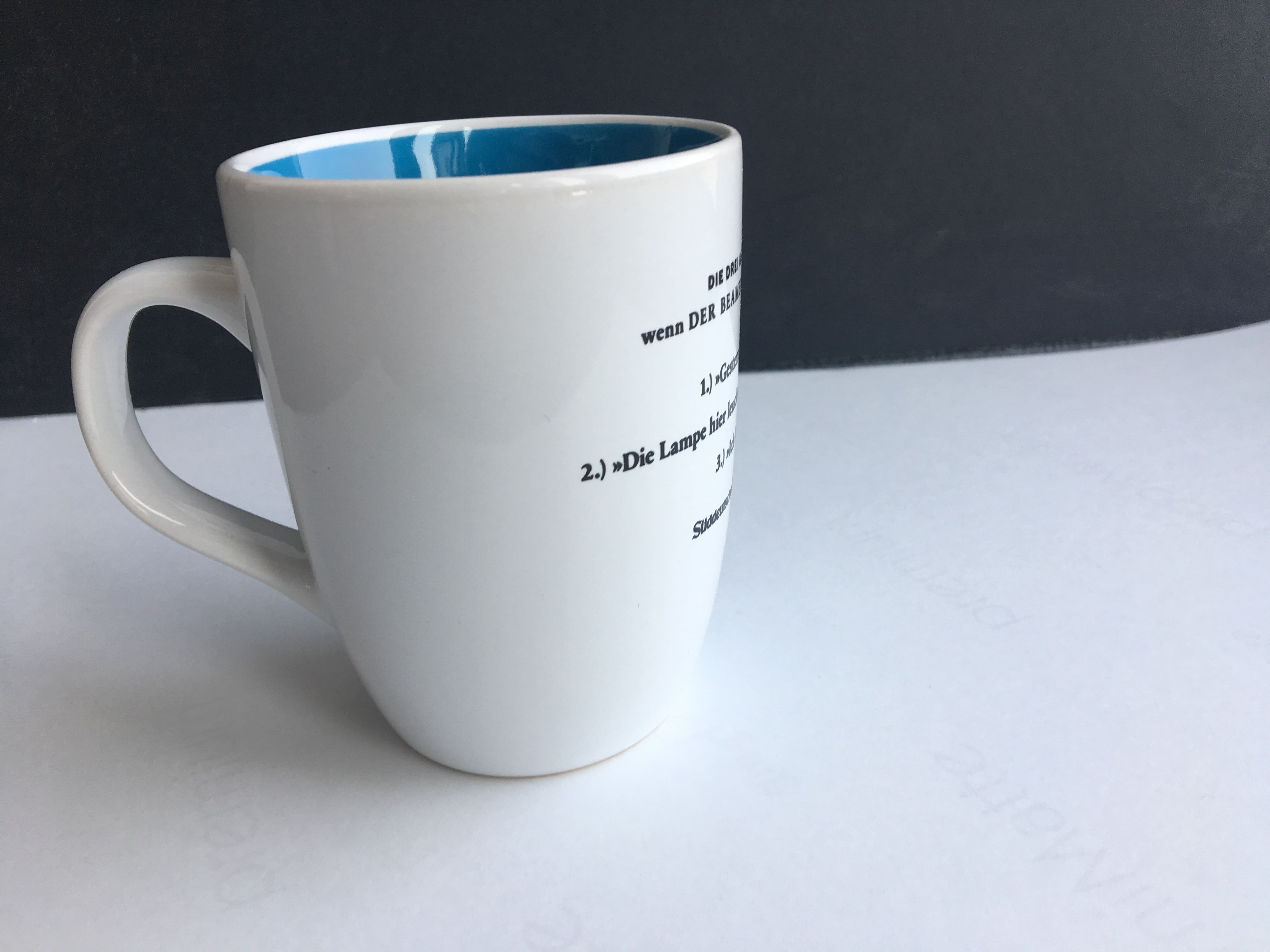 Weiße Keramiktasse mit schwarzem Text, blauer Innenseite, auf einer weißen Fläche vor einem dunklen Hintergrund.