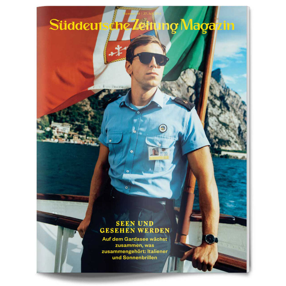 Ein Mann in blauer Uniform und mit Sonnenbrille steht auf einem Boot, mit Bergen und Fahnen im Hintergrund.
