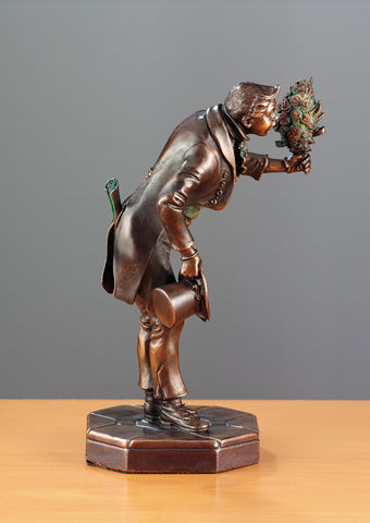 Bronzestatue eines Mannes im Anzug, der an einem Blumenstrauß riecht, einen Zylinder hält und auf einem sechseckigen Sockel steht.