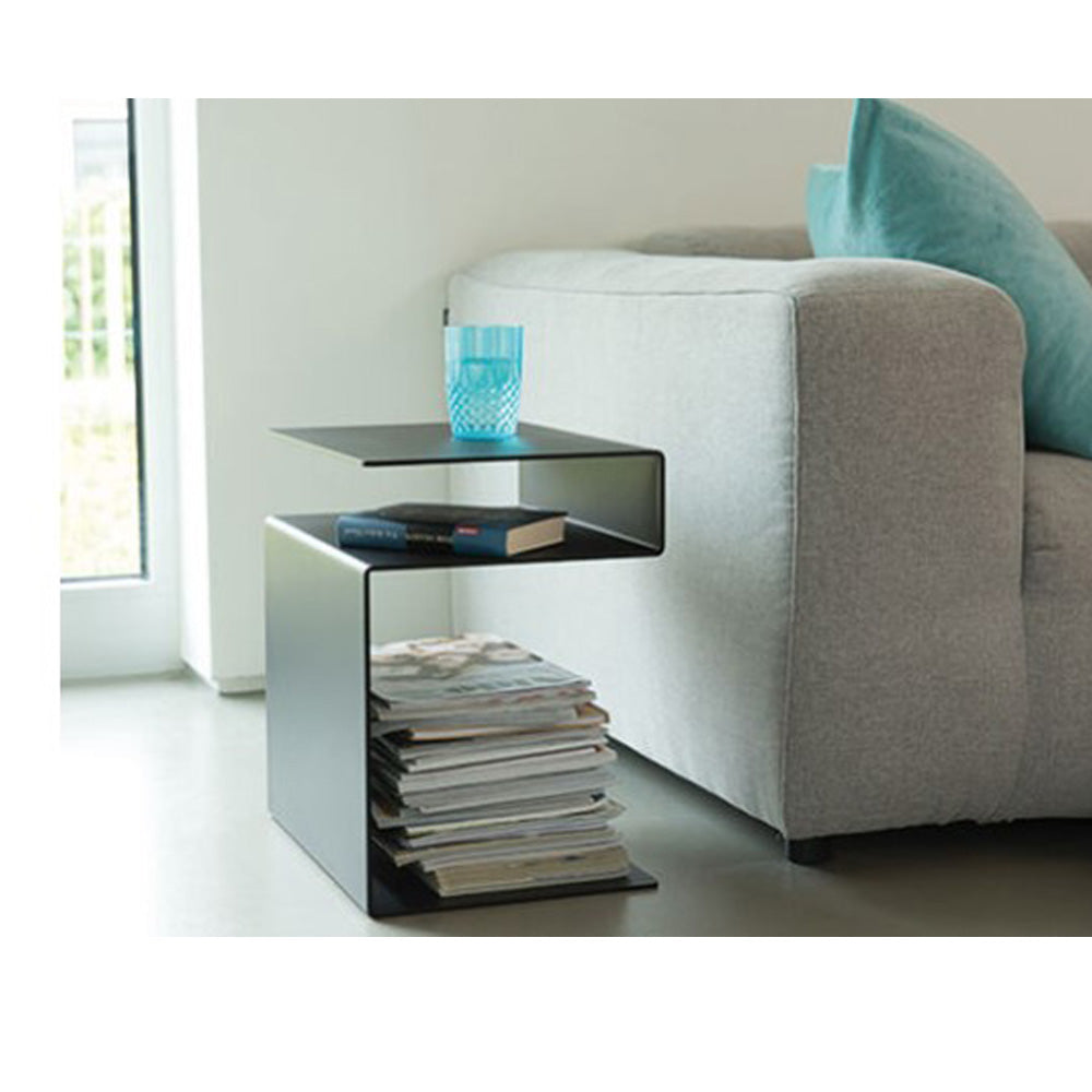Schwarzer moderner Beistelltisch neben einem grauen Sofa, mit einem Glas, Büchern und einem Stapel Zeitschriften.