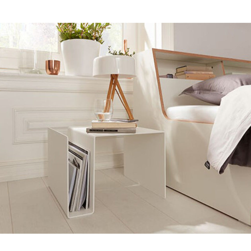 Moderner weißer Nachttisch mit Zeitschriften, Lampe, Pflanze und Glas Wasser neben einem Bett in einem hellen Schlafzimmer.