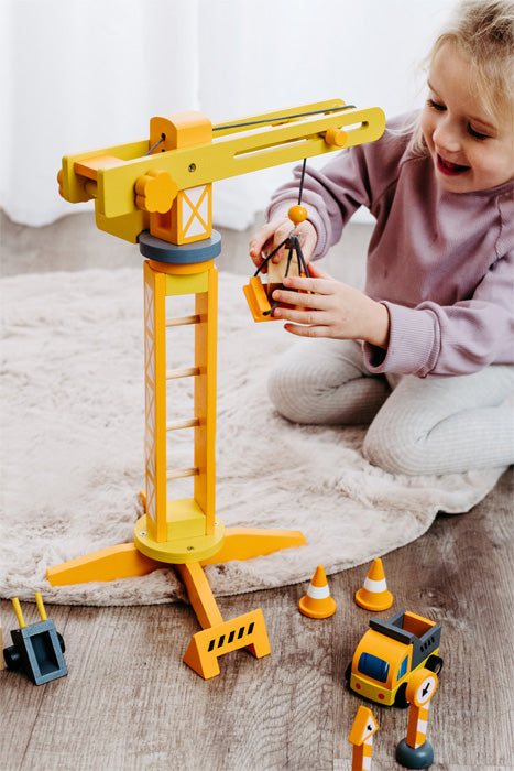 Ein lächelndes Kind spielt mit dem Baukran mit Baustellenzubehör, darunter ein gelber Spielzeugkran und Baufahrzeuge auf einem Holzboden.