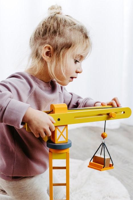 Ein junges Mädchen in einem hellvioletten Pullover spielt drinnen mit dem Baukran mit Baustellenzubehör, einem gelben Spielzeugkran-Set.