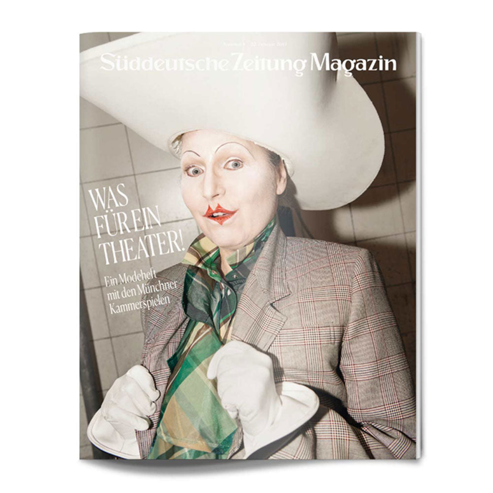 Person mit Theaterschminke, großem weißen Hut, karierter Jacke, Schal und Handschuhen auf einem Zeitschriftenumschlag mit deutschem Text.
