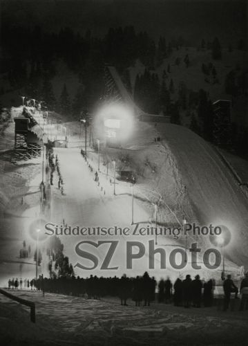Ein nächtliches Skispringen mit Zuschauern in der Nähe einer hell erleuchteten Skisprungschanze im Winter.