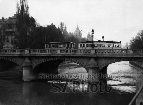 Historische Straßenbahn überquert eine Steinbrücke über einen Fluss mit Bäumen und Gebäuden im Hintergrund, in schwarz-weiß.