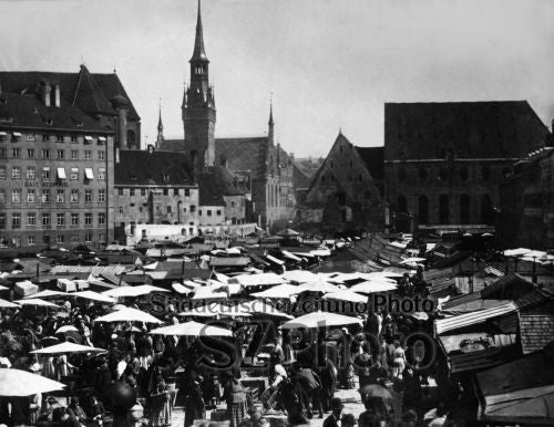 Historisches Schwarz-Weiß-Foto eines belebten Marktes auf einem europäischen Stadtplatz mit vielen Regenschirmen.