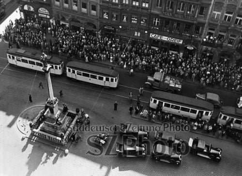 Luftaufnahme einer belebten Stadtstraße mit Straßenbahnen, Autos, einer Menschenmenge und einer Statue auf einem zentralen Platz.