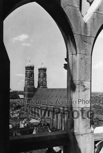 Blick auf die Zwillingstürme der Frauenkirche in München, umrahmt von einem steinernen Bogen, unter einem teilweise bewölkten Himmel.